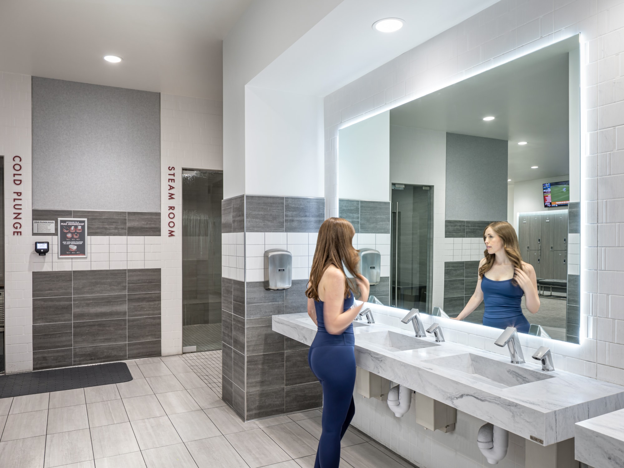 Modern commercial fitness facility restroom with marble sinks, lighted mirror, and hand dryers